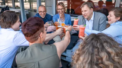 Ein Team stößt auf einer Terrasse mit bunten Getränken an; mehrere Kolleginnen und Kollegen lachen und halten ihre Gläser in die Mitte.