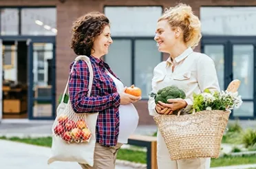 Zwei Frauen unterhalten sich lächelnd auf der Straße, eine hält Obst im Beutel, die andere Brokkoli und Blumen im Korb.
