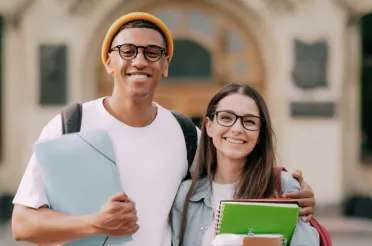 Ein Student und eine Studentin stehen lächelnd mit Mappen und Heften vor einem Universitätsgebäude.