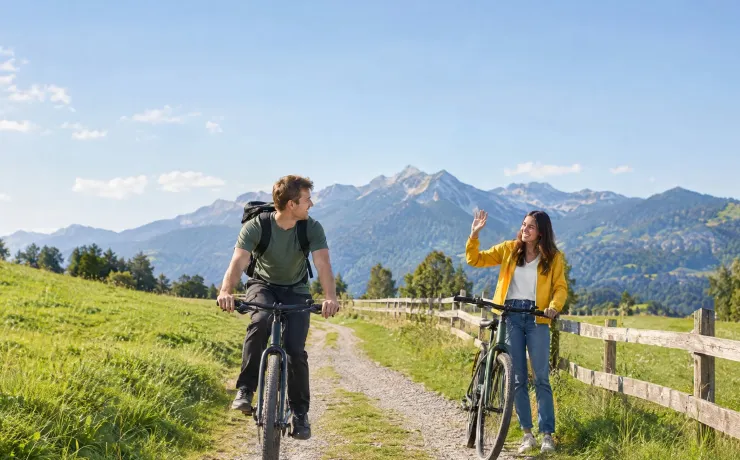 Ein junger Mann rollt auf dem Fahrrad über einen Feldweg zwischen Almwiesen auf seine winkende Freundin zu
