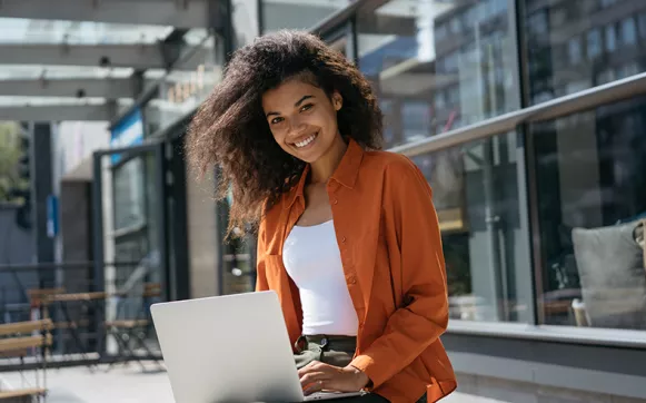 Marketing Studium Eine Frau sitzt draußen auf einer Bank mit Laptop und lacht in die Kamera