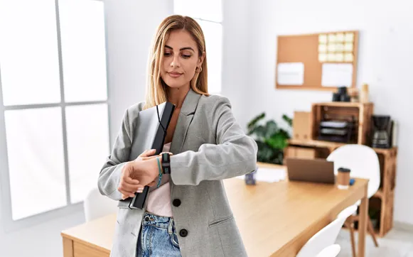 Eine Frau in Blazer schaut im Büro auf ihre Armbanduhr und hält einen Ordner unter dem Arm.
