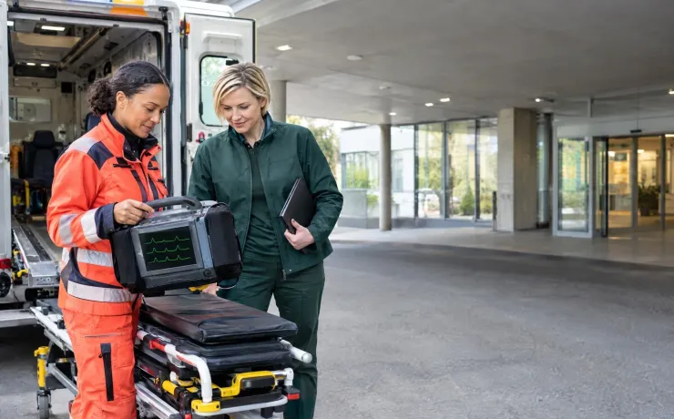 Notfallsanitäterin hält an der Liegendanfahrt den Monitor-Defibrillator, die Interoperabilitätsverantwortliche nickt, weil die vorausgeschickten Daten vollständig angekommen sind