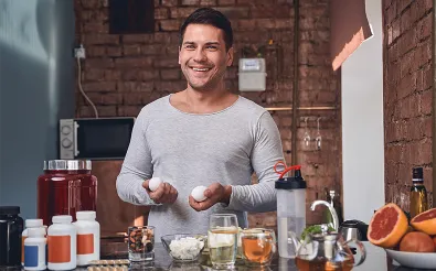 Ein lächelnder Mann steht in einer Küche mit Eiern in den Händen vor Proteinpulver, Dosen, Tee und Grapefruit.