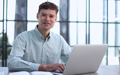 Ein junger Mann arbeitet vor einer großen Fensterfront an einem Laptop; neben ihm liegt ein aufgeschlagener Notizblock.