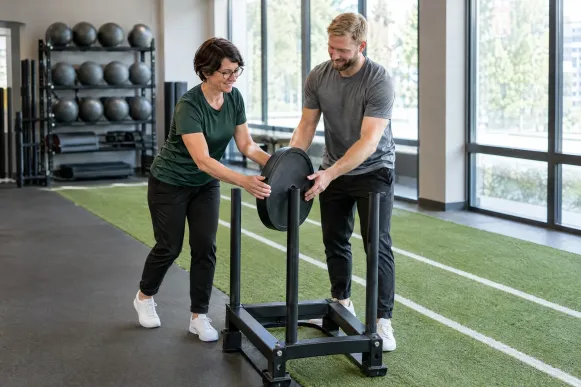 Studioleiterin und Trainer schieben im Fitnessstudio gemeinsam eine Gewichtsscheibe auf die Stange eines Trainingsschlittens