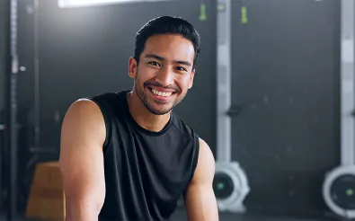 Ein lächelnder Mann in Sportshirt sitzt in einem Fitnessstudio und blickt freundlich in die Kamera.