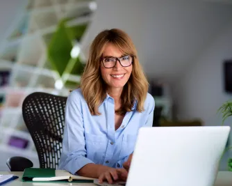 Eine Frau mit Brille sitzt am Schreibtisch vor einem Laptop und schaut lächelnd in die Kamera.