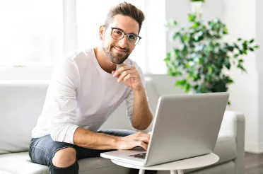 Ein Mann mit Brille sitzt lächelnd auf einem Sofa und arbeitet am Laptop.