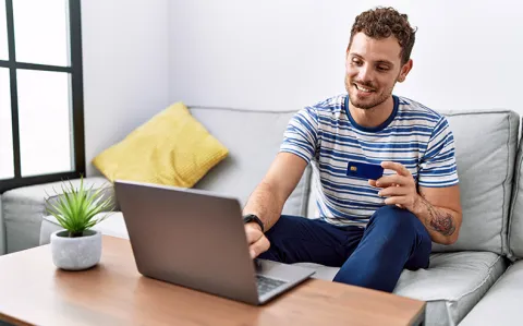 Ein Mann sitzt mit einer Bankkarte in der Hand auf dem Sofa vor einem Laptop auf dem Wohnzimmertisch.