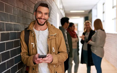 Ein junger Mann mit Mütze lehnt mit einem Smartphone in der Hand an einer Backsteinwand; im Hintergrund unterhalten sich vier Personen.