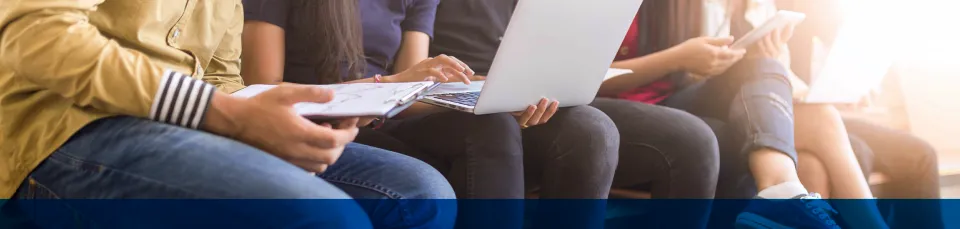 Eine Gruppe sitzt nebeneinander, Laptops und Unterlagen auf den Knien, unten blau abgesetzter Bildrand.