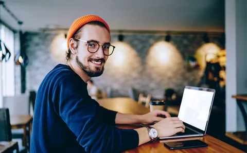 Ein junger Mann mit oranger Mütze arbeitet in einem Café am Laptop und schaut lächelnd über die Schulter in die Kamera.