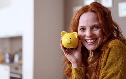 Eine lächelnde Frau hält ein gelbes Sparschwein an ihre Wange und steht für die Finanzierung des Studiums.
