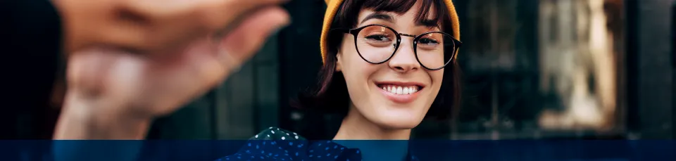 Eine lächelnde Frau mit Brille und gelber Mütze blickt in die Kamera, im Vordergrund greift eine Hand ins Bild.
