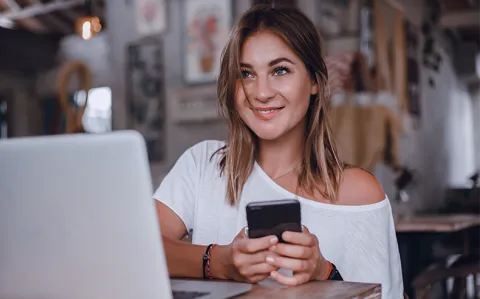 Eine Frau sitzt lächelnd mit einem Smartphone in der Hand vor einem Laptop in einem Café.