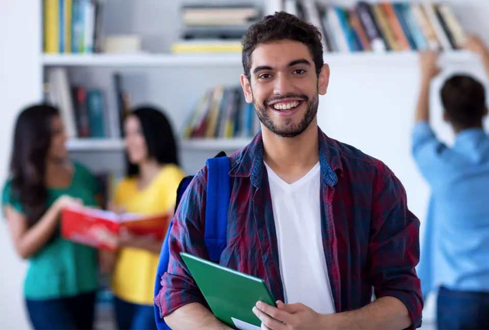 Student mit Buch lacht in die Kamera. Im Hintergrund lernen weitere Studenten.