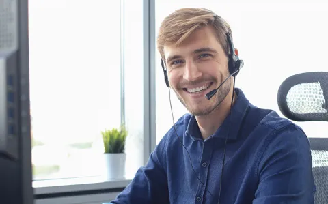 Ein Mitarbeiter der Studienberatung mit Headset sitzt am Arbeitsplatz und schaut lächelnd in die Kamera.