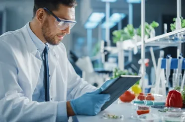 Ein Mann in Laborkittel und Schutzbrille liest in einem Labor auf einem Tablet.