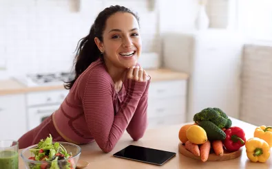 Eine lächelnde Frau stützt sich in einer Küche auf den Tisch, davor liegen Gemüse, Salat und ein Tablet.