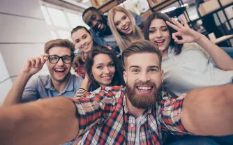 Eine Gruppe junger Menschen drängt sich lachend für ein Selfie zusammen; eine Person zeigt das OK-Zeichen.