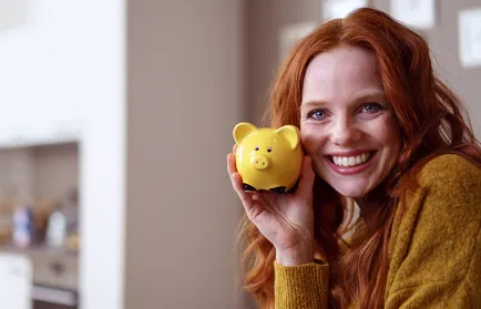 Eine lächelnde Frau hält ein gelbes Sparschwein an ihre Wange und steht für die Finanzierung des Studiums.