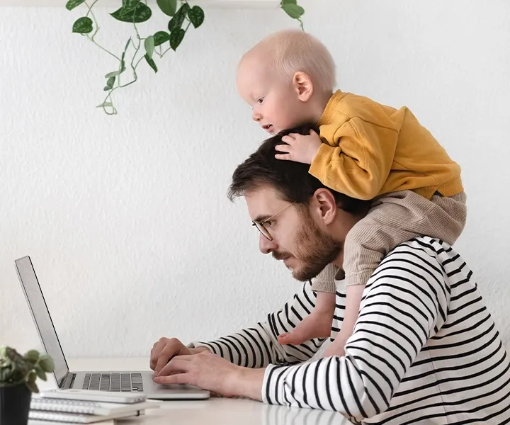 Ein Vater arbeitet am Laptop, während sein kleines Kind auf seinen Schultern sitzt.