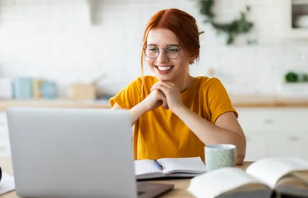 Eine lächelnde Frau mit Brille sitzt am Laptop, vor ihr liegen aufgeschlagene Bücher und eine Tasse.