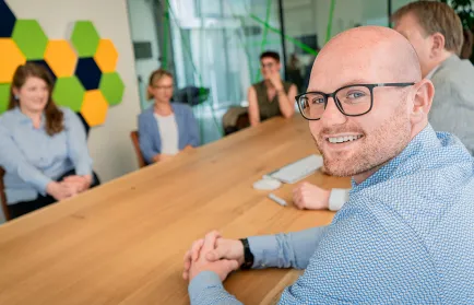 Ein Mann mit Brille schaut in einer Besprechung über die Schulter in die Kamera; im Hintergrund vier Kolleginnen und Kollegen am Konferenztisch.