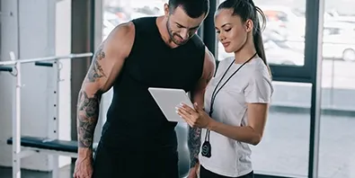 Ein Trainer und eine Trainerin besprechen in einem Fitnessstudio gemeinsam Inhalte auf einem Tablet.