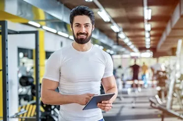 Ein lächelnder Mann mit Bart steht mit einem Tablet in einem Fitnessstudio.