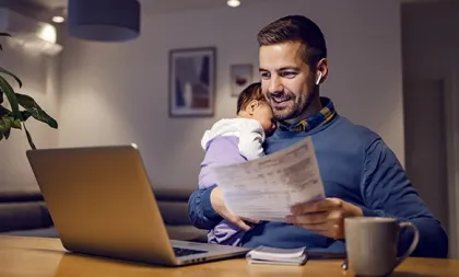 Ein Vater hält ein Baby auf dem Arm und liest lächelnd ein Schreiben mit Zahlen; vor ihm stehen ein Laptop und eine Kaffeetasse.