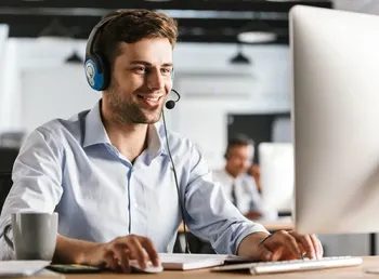 Ein Mann mit Headset arbeitet lächelnd am Computer, im Hintergrund sitzt ein Kollege.