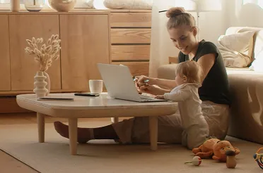 Eine Frau sitzt mit ihrem Kleinkind auf dem Boden und arbeitet am Laptop auf dem Couchtisch.