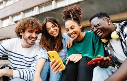 Vier junge Menschen sitzen dicht nebeneinander auf einer Treppe vor einem Gebäude, schauen gemeinsam auf ein Smartphone und lachen.