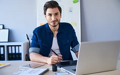Ein junger Mann sitzt mit Grafiktablett und Stift vor einem Laptop; im Hintergrund eine Wand mit Entwurfsskizzen.