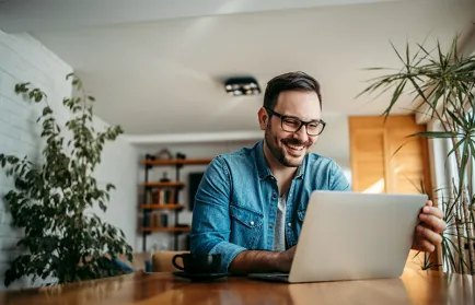 Ein Mann mit Brille sitzt lachend an einem Holztisch vor einem Laptop; neben ihm eine Kaffeetasse, im Raum mehrere Zimmerpflanzen.