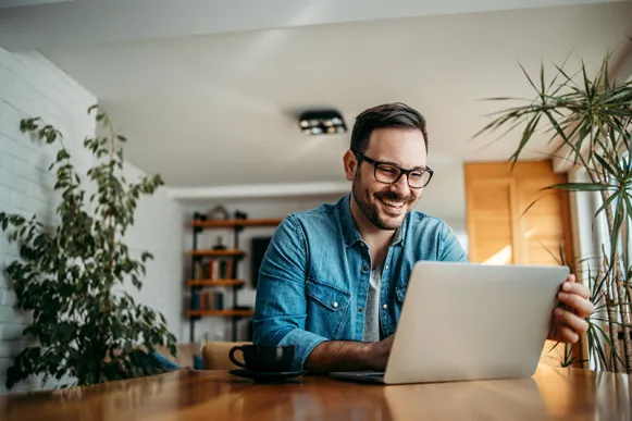 Ein Mann mit Brille sitzt lachend an einem Holztisch vor einem Laptop; neben ihm eine Kaffeetasse, im Raum mehrere Zimmerpflanzen.