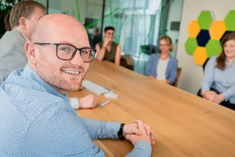 Ein lächelnder Mann mit Brille sitzt an einem langen Holztisch, im Hintergrund besprechen sich Kollegen.