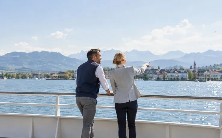 Ein Mann in Arbeitsweste und eine Frau im Blazer blicken an der Reling einer Bodensee-Fähre gemeinsam dem nahenden Ufer entgegen