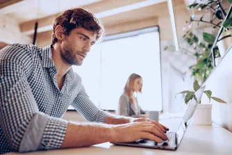 Ein Mann im karierten Hemd tippt konzentriert auf seinem Laptop in einem hellen Büro; im Hintergrund arbeitet eine Kollegin.