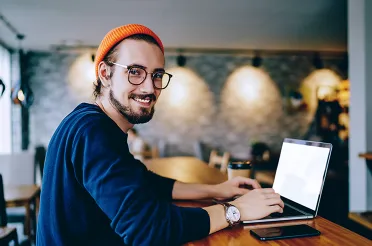 Ein junger Mann mit oranger Mütze arbeitet in einem Café am Laptop und schaut lächelnd über die Schulter in die Kamera.