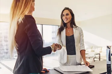 Zwei Frauen geben sich in einem hellen Büro lächelnd die Hand, auf dem Tisch liegen Unterlagen.