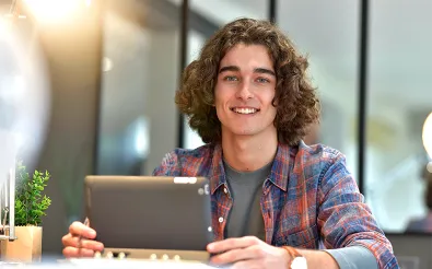 Ein junger Mann mit lockigem Haar sitzt an einem Tisch hinter einem Tablet und schaut lächelnd in die Kamera.