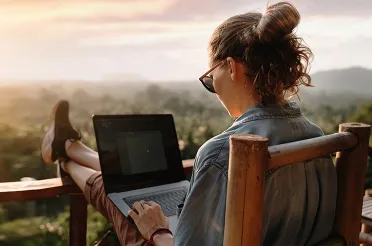 Eine Frau sitzt mit hochgelegten Beinen auf einer Terrasse über einer Berglandschaft und arbeitet am Laptop.