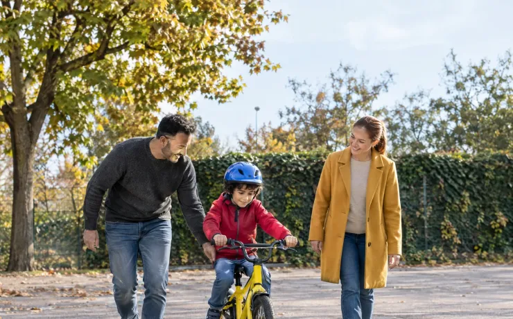 Ein Kind fährt auf einem Schulparkplatz zum ersten Mal ohne Stützräder, der Vater hält den Sattel und die Mutter schaut stolz zu