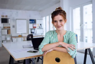 Frau sitzt im Büro & lacht in die Kamera. Laptop und Handy liegen auf dem Tisch.