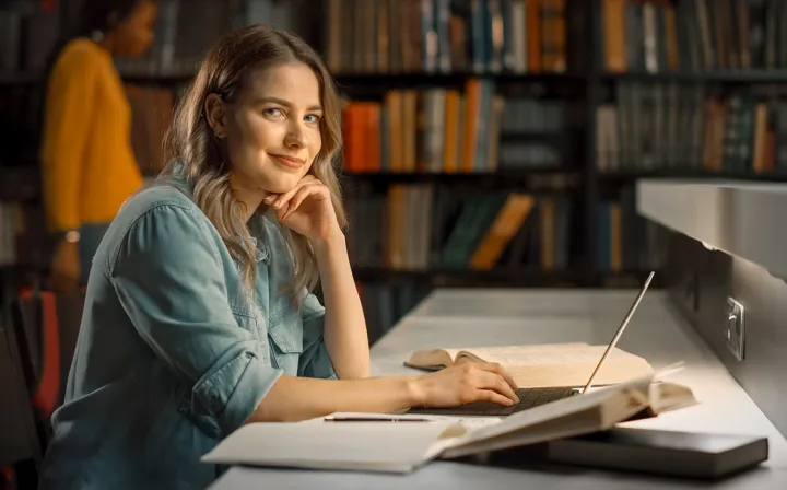 Eine Studentin stützt in einer Bibliothek lächelnd das Kinn auf die Hand, vor ihr liegen Laptop und Bücher.