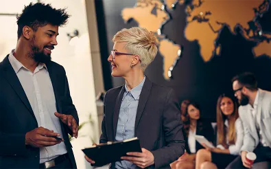 Ein Mann und eine Frau im Business-Look unterhalten sich lächelnd vor einer Weltkarte an der Wand, dahinter sitzen Kolleginnen und Kollegen.