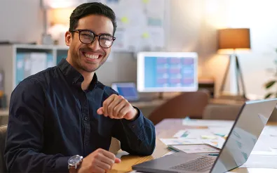 Ein lächelnder Mann mit Brille sitzt an einem Schreibtisch mit Laptop und Unterlagen im Büro.